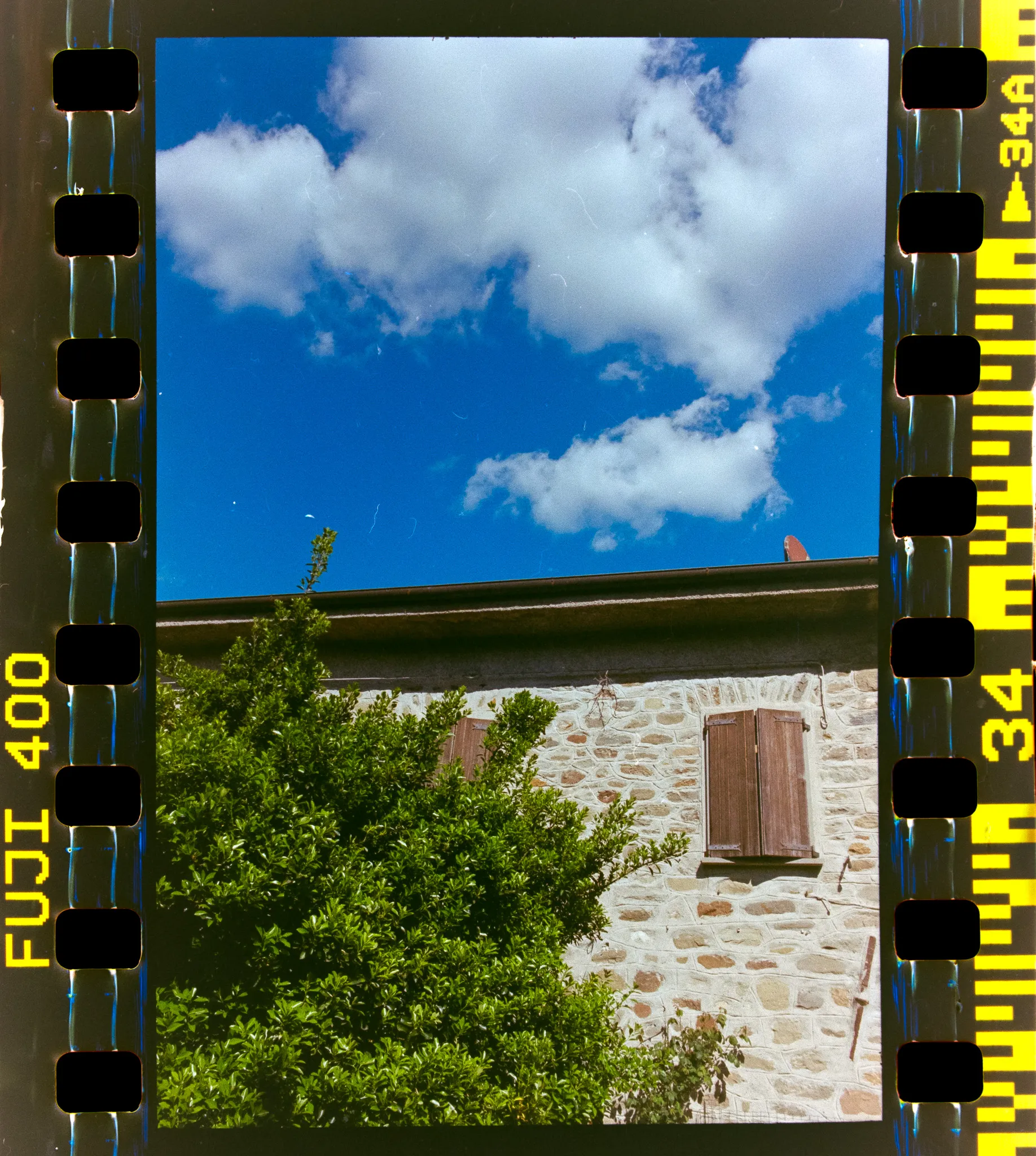 Casa in pietra con persiane e rampicante sotto un cielo blu con nuvole, pellicola a colori